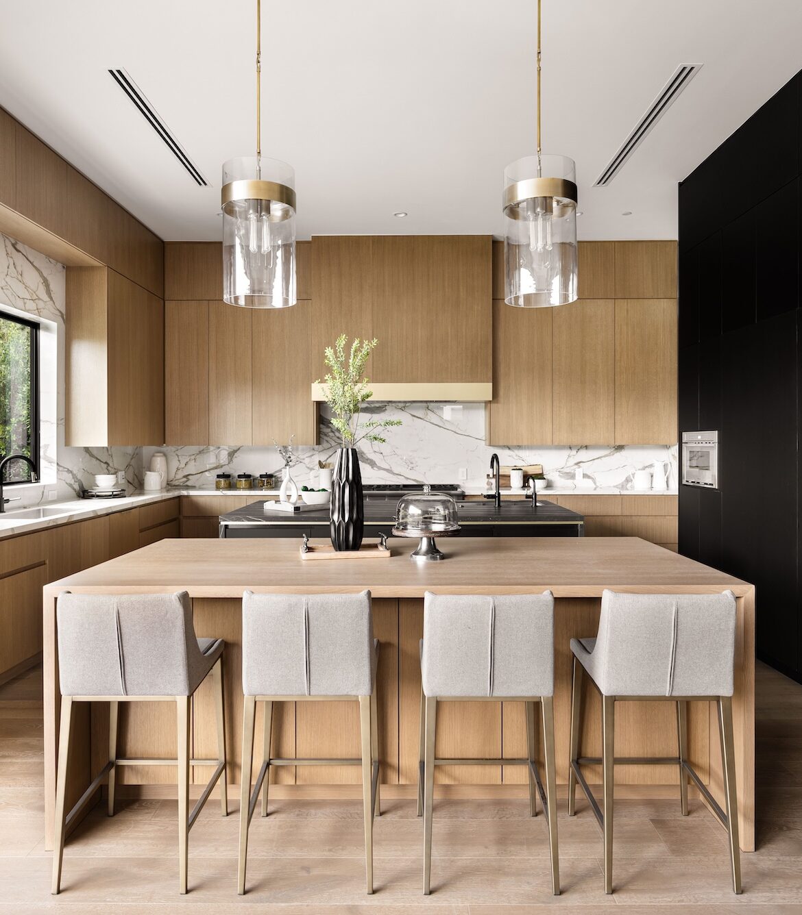 Modern kitchen interior with wooden cabinets, marble countertops, and a central island with bar stools under pendant lighting.